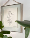 Die Seepferdchenkarte hängt in einem gläsernen Bilderramen an der Wand. Im Vordergrund sind grüne Blätter zu erkennen.